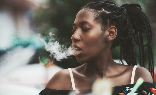 Portrait of a dazzling African girl with braids sitting in a street cafe and exhaling smoke from the hookah; young black female outdoors in a bar plays with the vapor from an electronic cigarette